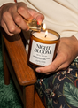 Person lighting a 'Night Bloom' candle with a wooden chair and floral-patterned fabric in the background.