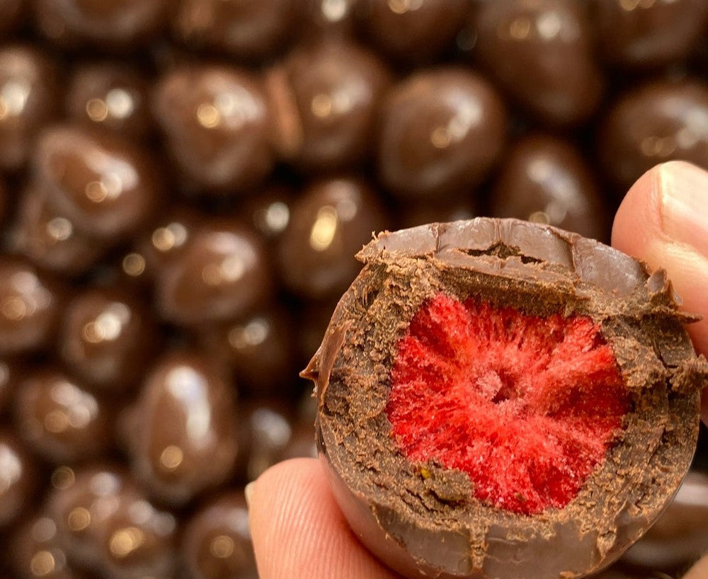 Whole strawberry that has been freeze dried and covered with chocolate being held with two fingers and has had a bite take out of it to reveal the strawberry within the chocolate.