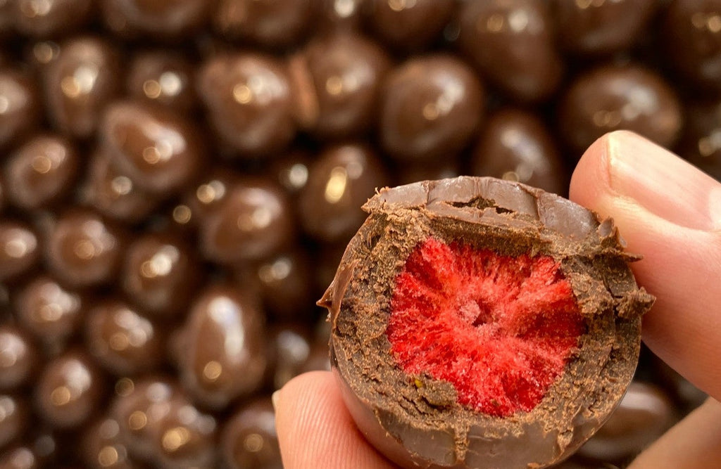 Whole strawberry that has been freeze dried and covered with chocolate being held with two fingers and has had a bite take out of it to reveal the strawberry within the chocolate.